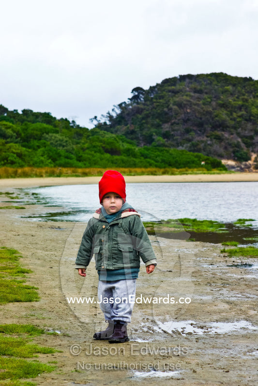 A small boy dressed in winter clothes walking along a river shore. Tidal River, Wilsons Promontory National Park, Victoria, Australia