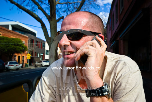 A balding man talks on his mobile phone whilst sitting at a cafe. Collingwood, Victoria, Australia