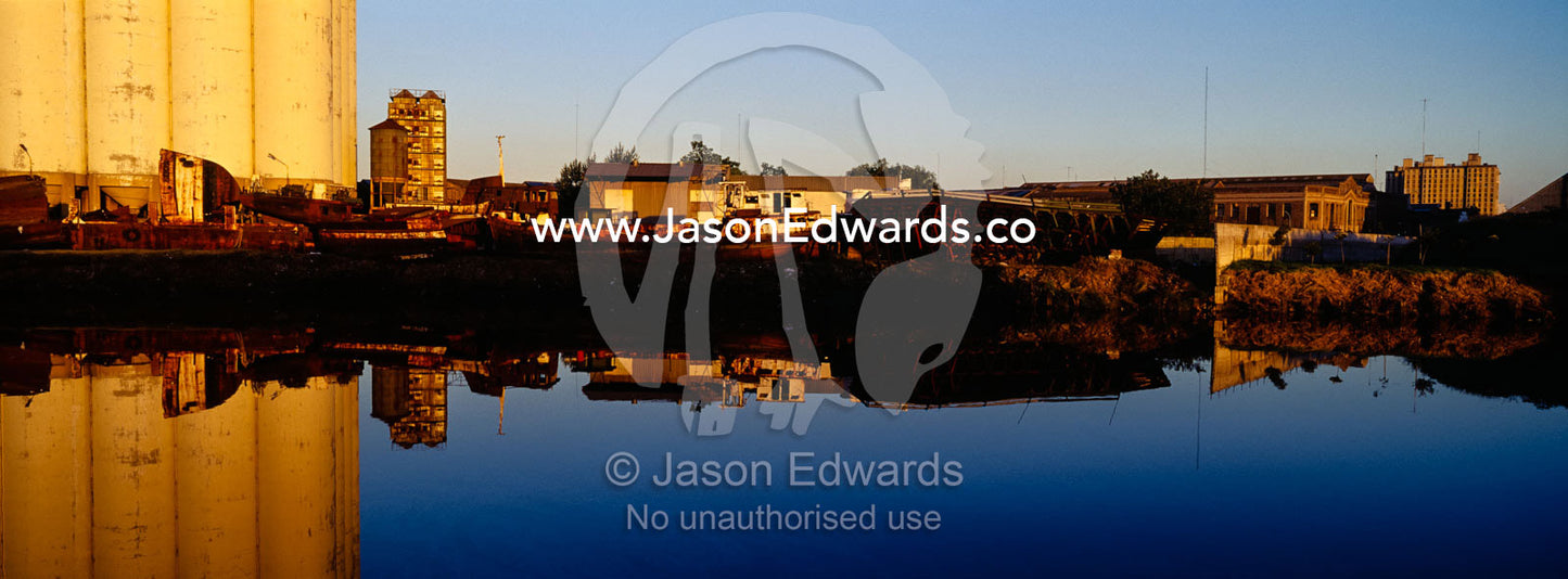 Grain silos and rusting boats reflect in the still waters of a river. Riachuelo River, La Boca, Buenos Aires, Argentina.