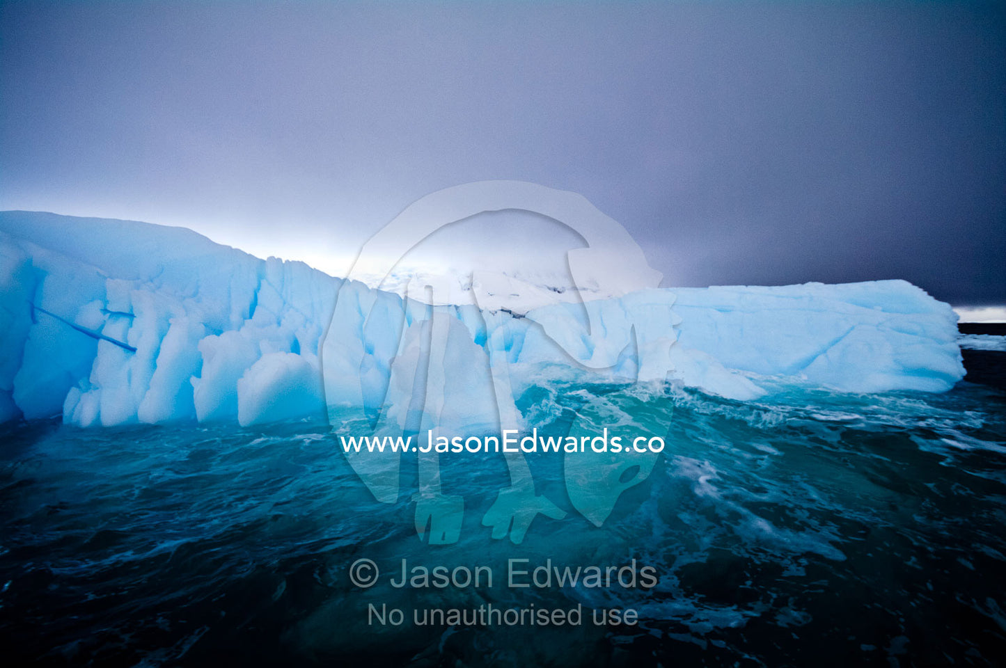 Waves wash against a deep blue iceberg beneath a stormy Antarctic sky. Crystal Sound, Hanusse Bay, Antarctic Peninsula, Antarctic Circle, Antarctica.