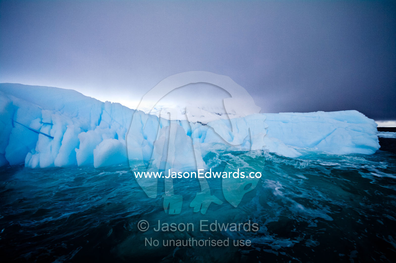 Waves wash against a deep blue iceberg beneath a stormy Antarctic sky. Crystal Sound, Hanusse Bay, Antarctic Peninsula, Antarctic Circle, Antarctica.