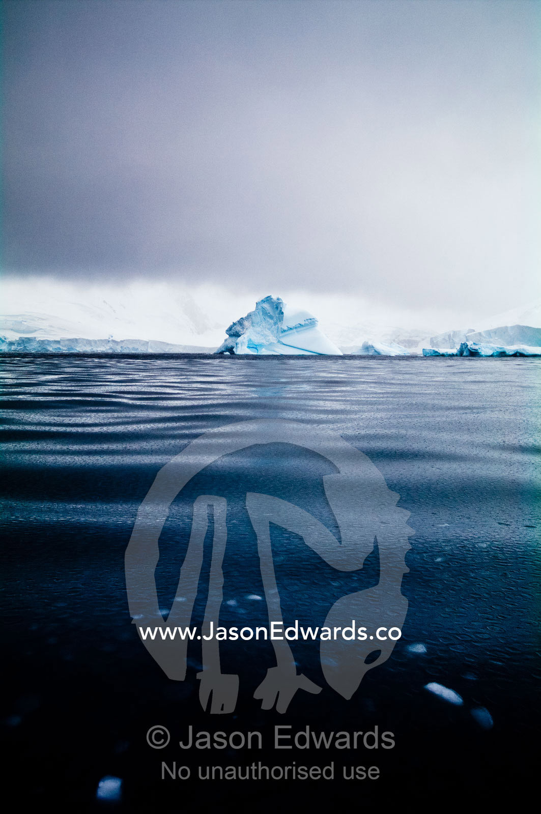 Under stormy skies an iceberg floats through mercury-like Frizzle Ice. Crystal Sound, Hanusse Bay, Antarctic Peninsula, Antarctic Circle, Antarctica.