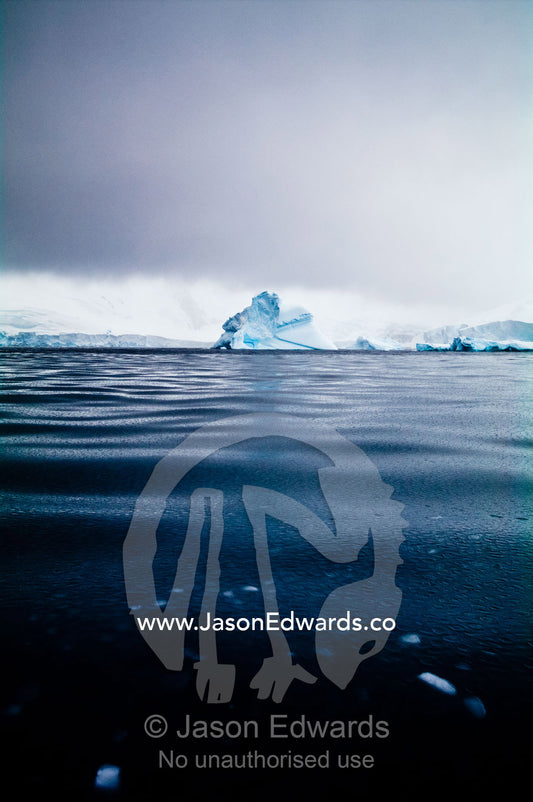 Under stormy skies an iceberg floats through mercury-like Frizzle Ice. Crystal Sound, Hanusse Bay, Antarctic Peninsula, Antarctic Circle, Antarctica.