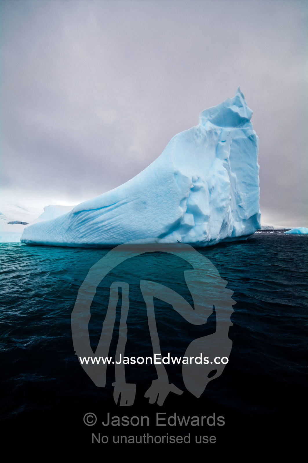 A pointy blue iceberg emerges from a dark sea beneath stormy skies. Crystal Sound, Hanusse Bay, Antarctic Peninsula, Antarctic Circle, Antarctica.