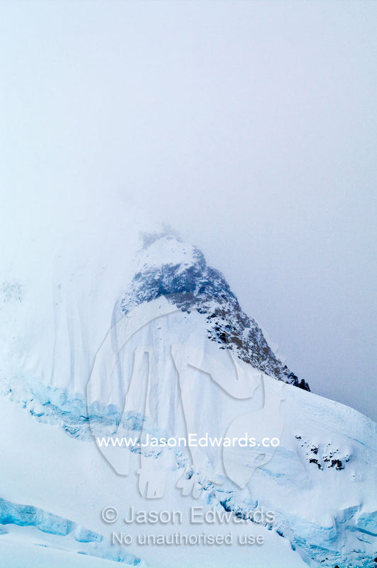 A jagged snow and ice encrusted mountain peak emerges from a storm. Crystal Sound, Hanusse Bay, Antarctic Peninsula, Antarctic Circle, Antarctica.