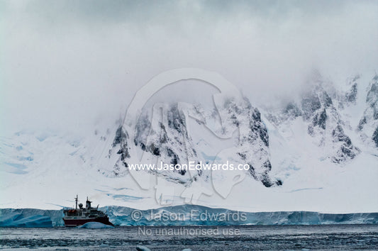 Vast and menacing mountains dwarf a glacier and ecotourism ship. Crystal Sound, Hanusse Bay, Antarctic Peninsula, Antarctic Circle, Antarctica.
