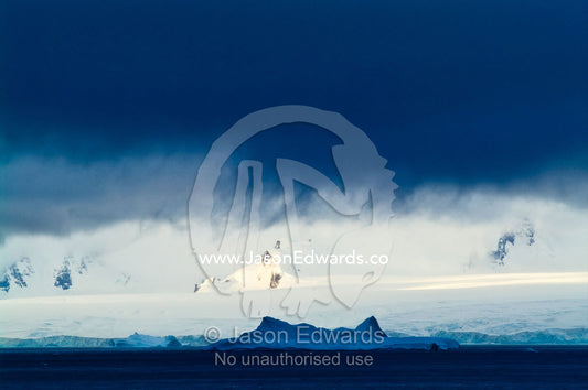 A dense storm descends over a mountain range dwarfing a large iceberg. Crystal Sound, Hanusse Bay, Antarctic Peninsula, Antarctic Circle, Antarctica.