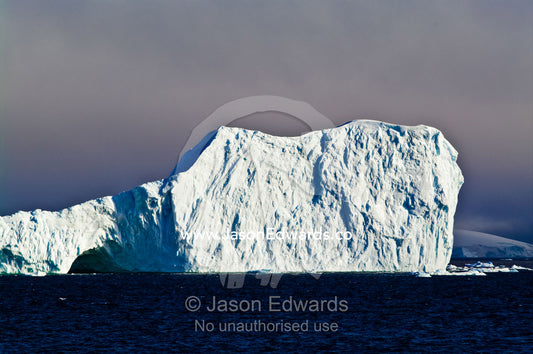 Sunlight bounces off the sheer rugged walls of an enormous iceberg. Fish Islands, Crystal Sound, Antarctic Peninsula, Antarctic Circle, Antarctica.