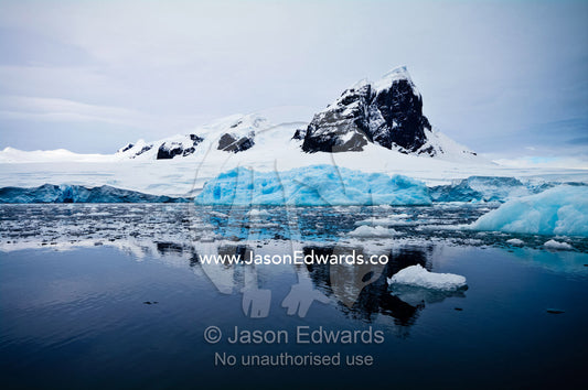 A sheer black mountain summit covered in snow and ice above icebergs. Fish Islands, Flouder Island, The Minnows, Crystal Sound, Antarctic Peninsula, Antarctic Circle, Antarctica.