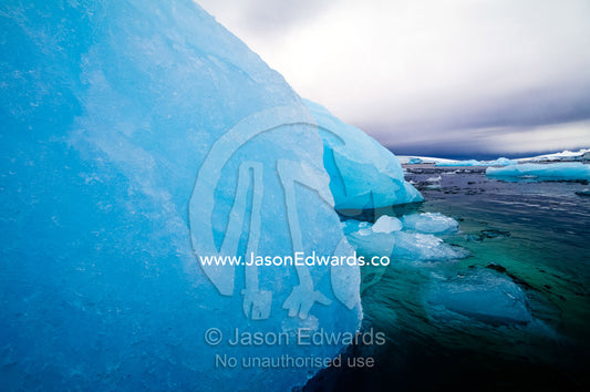 A ghostly grey storm washes over an iridescent blue iceberg in a bay. Fish Islands, Flouder Island, The Minnows, Crystal Sound, Antarctic Peninsula, Antarctic Circle, Antarctica.