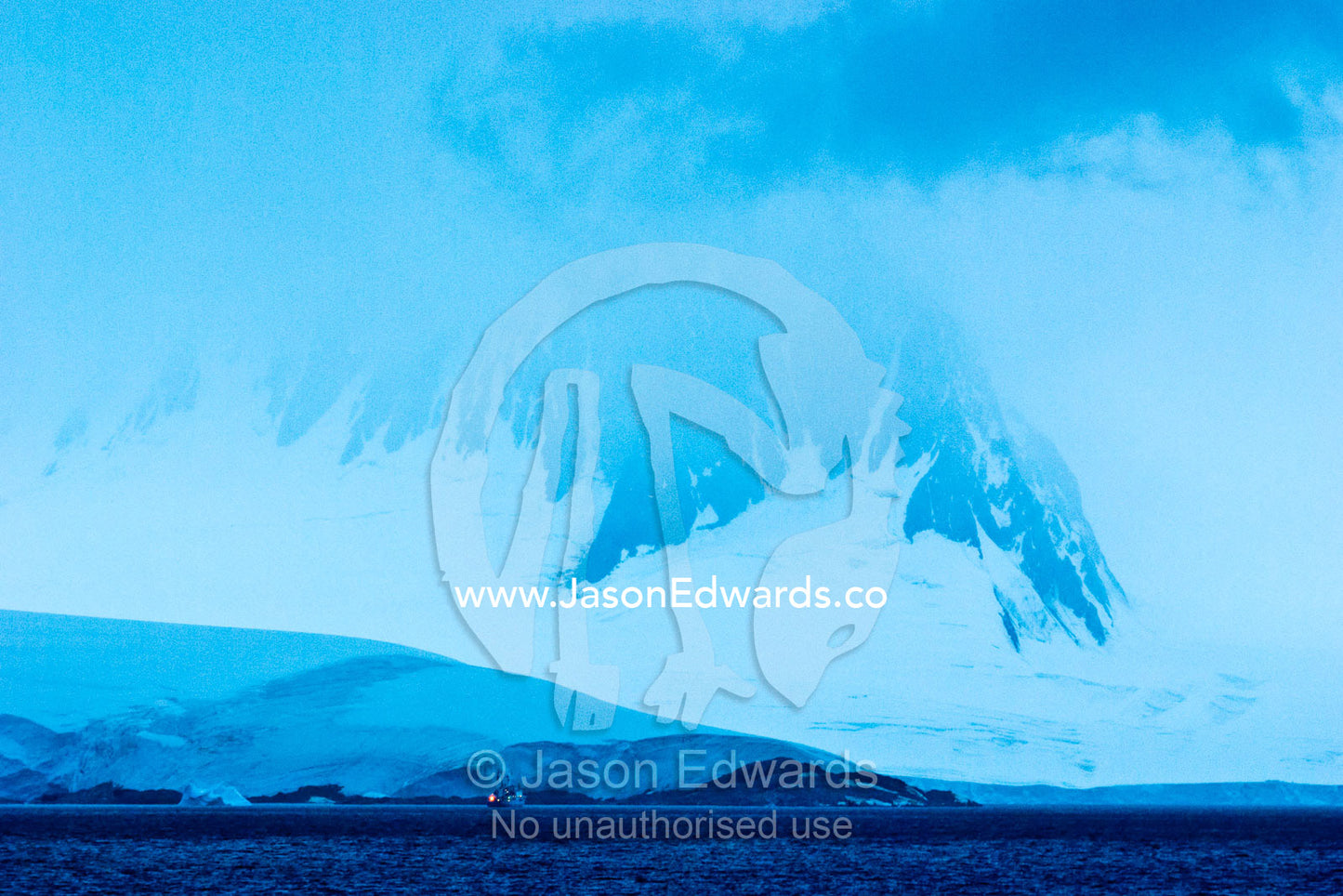 An Antarctic ship dwarfed by a glacier covered mountain range at night Port Lockroy, Gerlache Strait, Antarctic Peninsula, Antarctica.