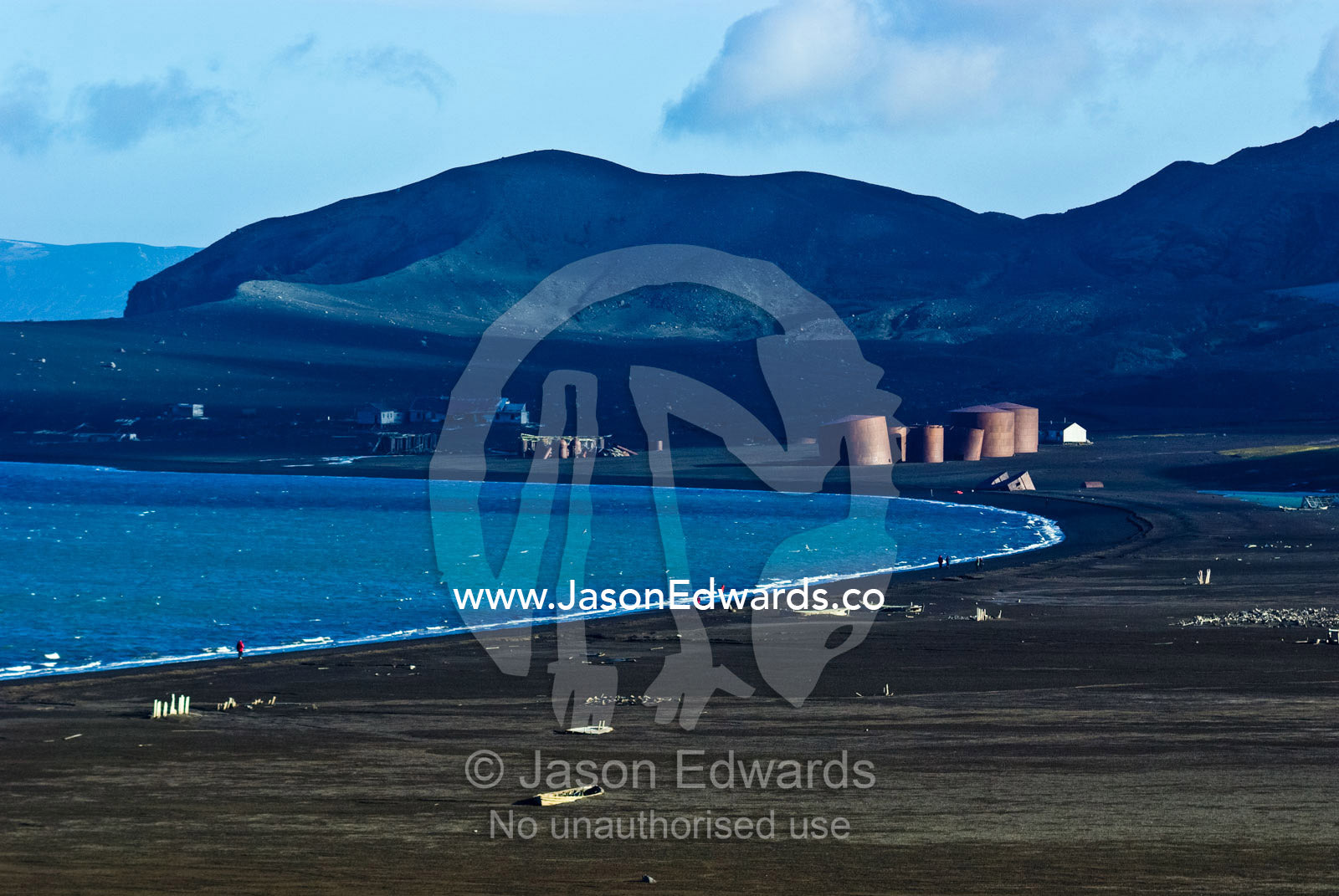 Tourists hike on a beach near abandoned whaling station oil tanks. Hektor Whaling Station, Whalers Bay, Port Foster, Deception Island, South Shetland Islands, Antarctic Peninsula, Antarctica.