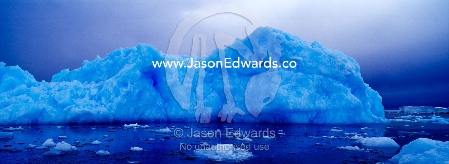 A jagged iridescent blue iceberg rises from a moody calm ocean. Fish Islands, The Minnows, Flounder Island, Crystal Sound, Antarctic Peninsula, Antarctic Circle, Antarctica.