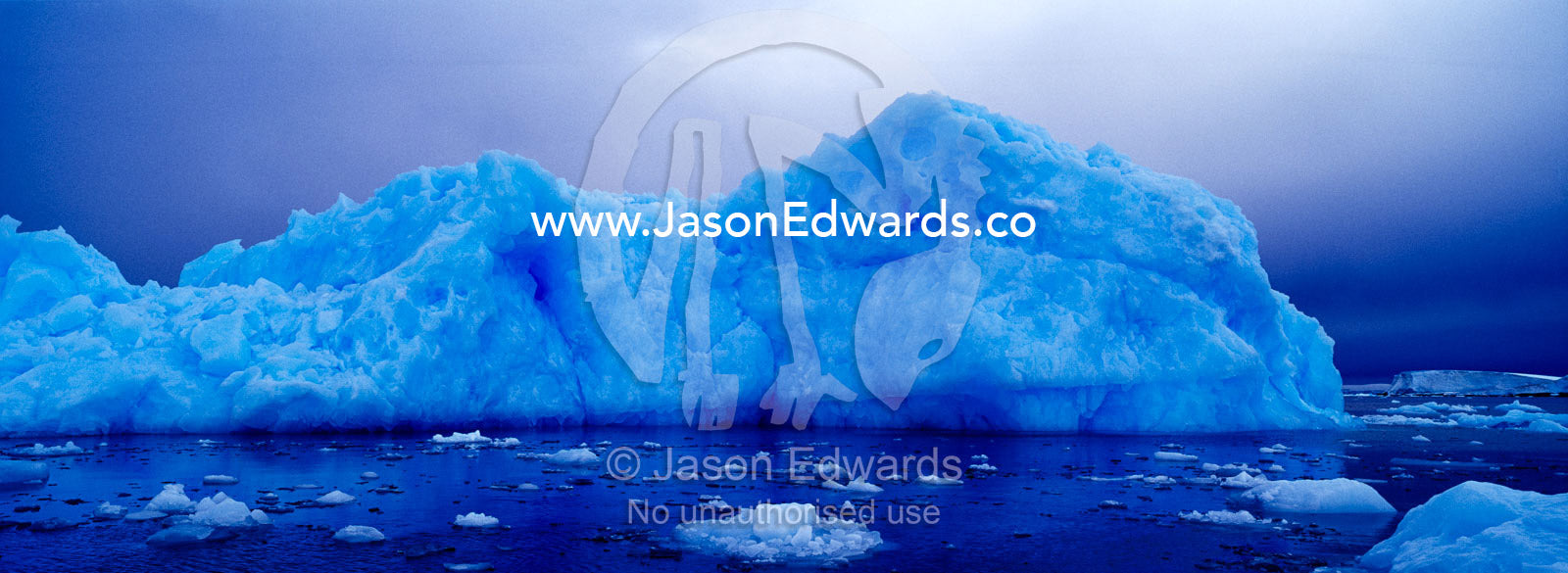 A jagged iridescent blue iceberg rises from a moody calm ocean. Fish Islands, The Minnows, Flounder Island, Crystal Sound, Antarctic Peninsula, Antarctic Circle, Antarctica.
