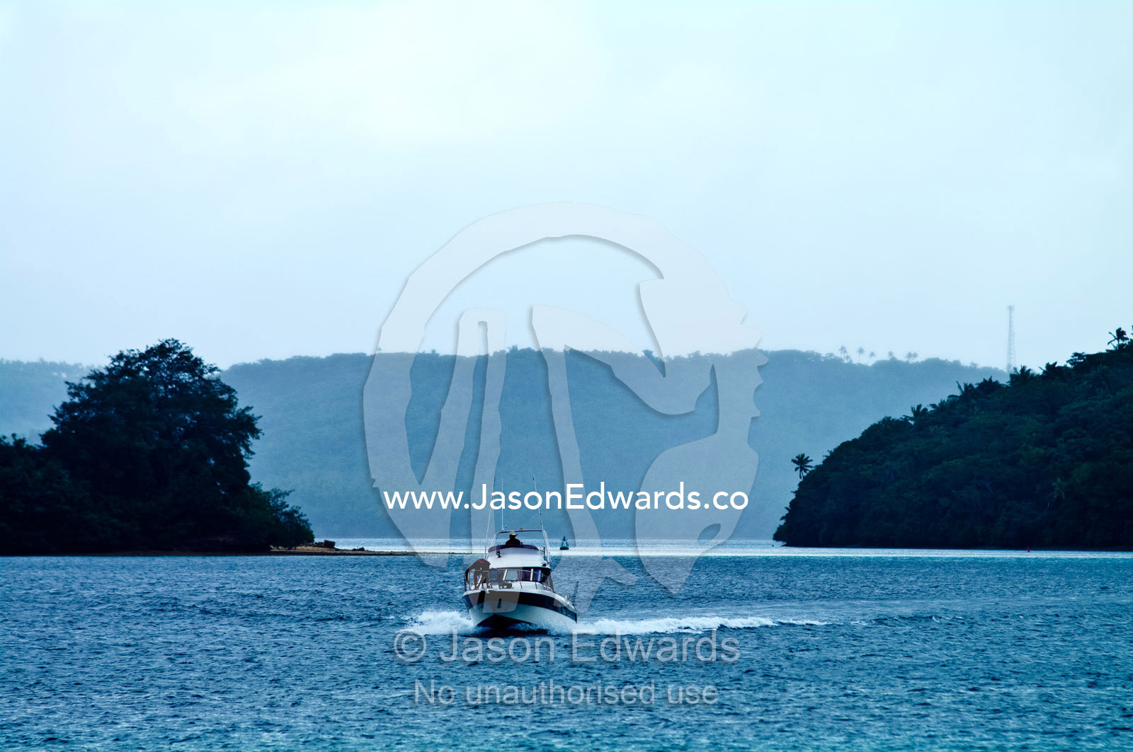 A motor boat speeds between two tropical islands in a small bay. Port of Refuge, Neiafu, Vava'u Island Group, Pacific Ocean, Oceania, Kingdom of Tonga.