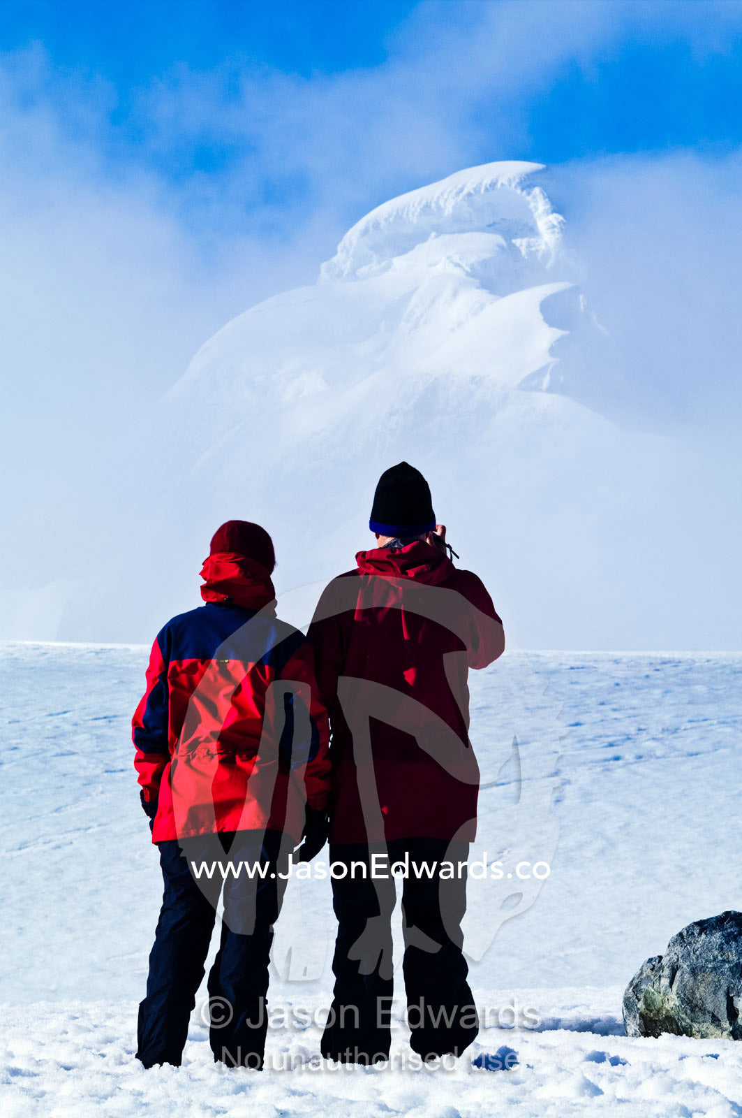 A tourist couple video an enormous ice mountain on a rugged island. Prospect Point, Crystal Sound, Antarctic Peninsula, Antarctic Circle, Antarctica.