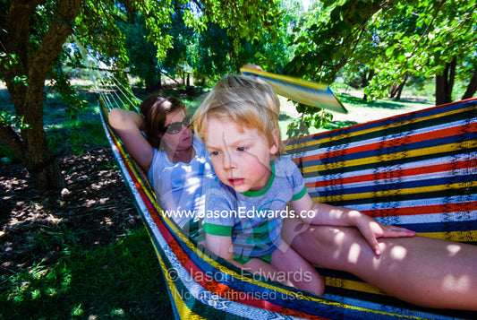Mother and son swing in a hammock under fruit trees on a summers day. Castlemaine, Victoria, Australia.