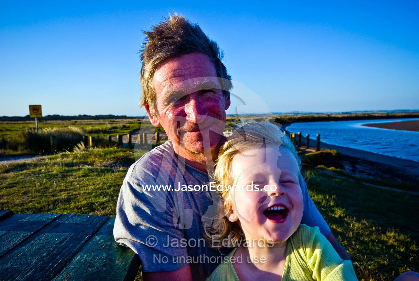 A father holds his laughing son by a river on a windy afternoon. Anderson's Inlet, Venus Bay, South Gippsland, Victoria, Australia.