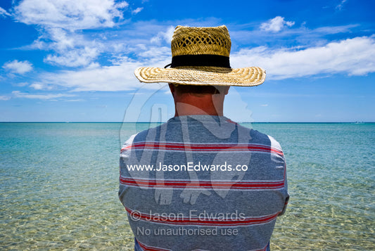 A man in a straw sun hat watches the ocean from the beach shallows. Rye Beach, Melbourne, Victoria, Australia.