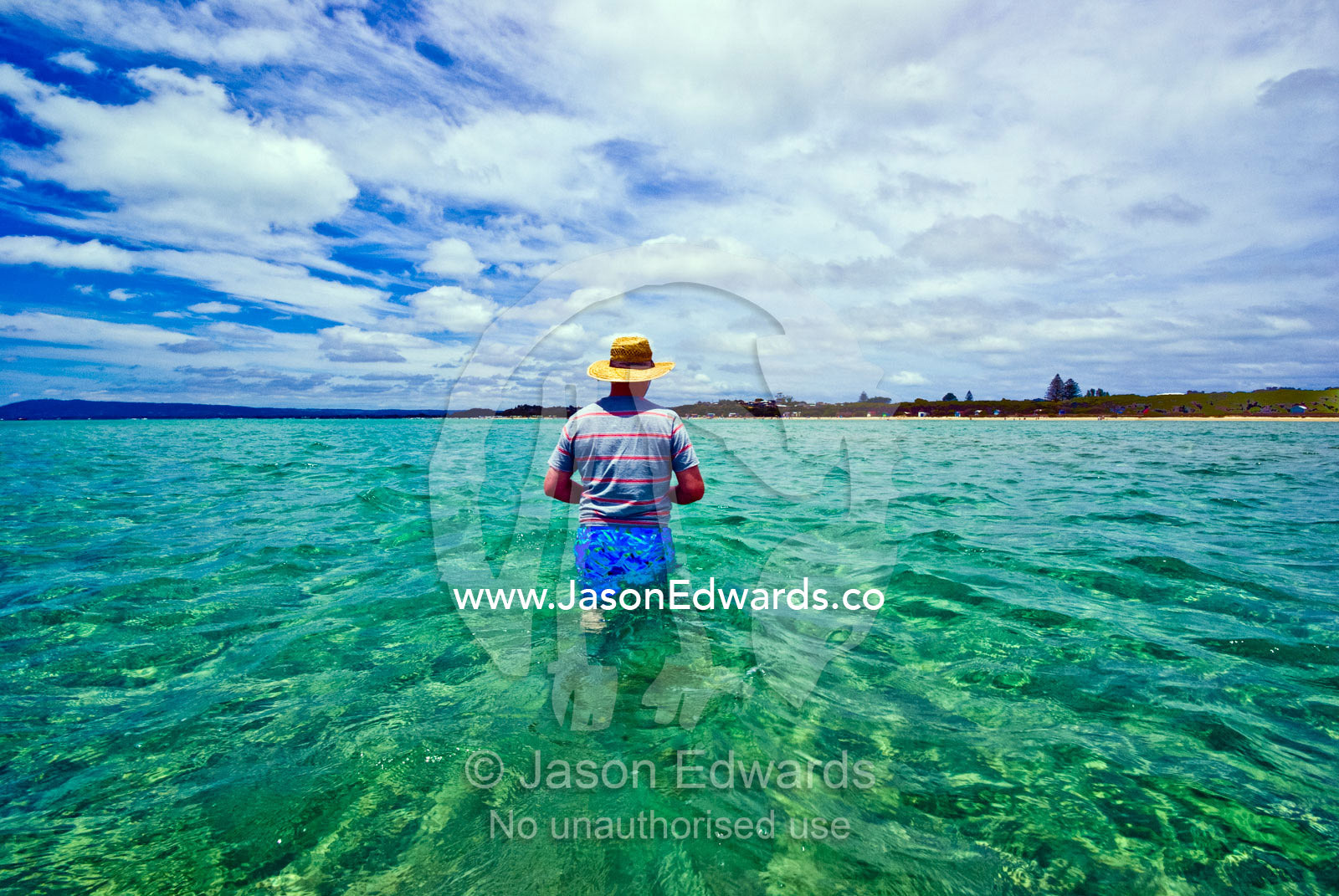 A man in a straw sun hat wades the ocean shallows towards the beach. Rye Beach, Melbourne, Victoria, Australia.