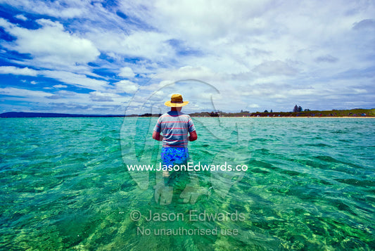A man in a straw sun hat wades the ocean shallows towards the beach. Rye Beach, Melbourne, Victoria, Australia.