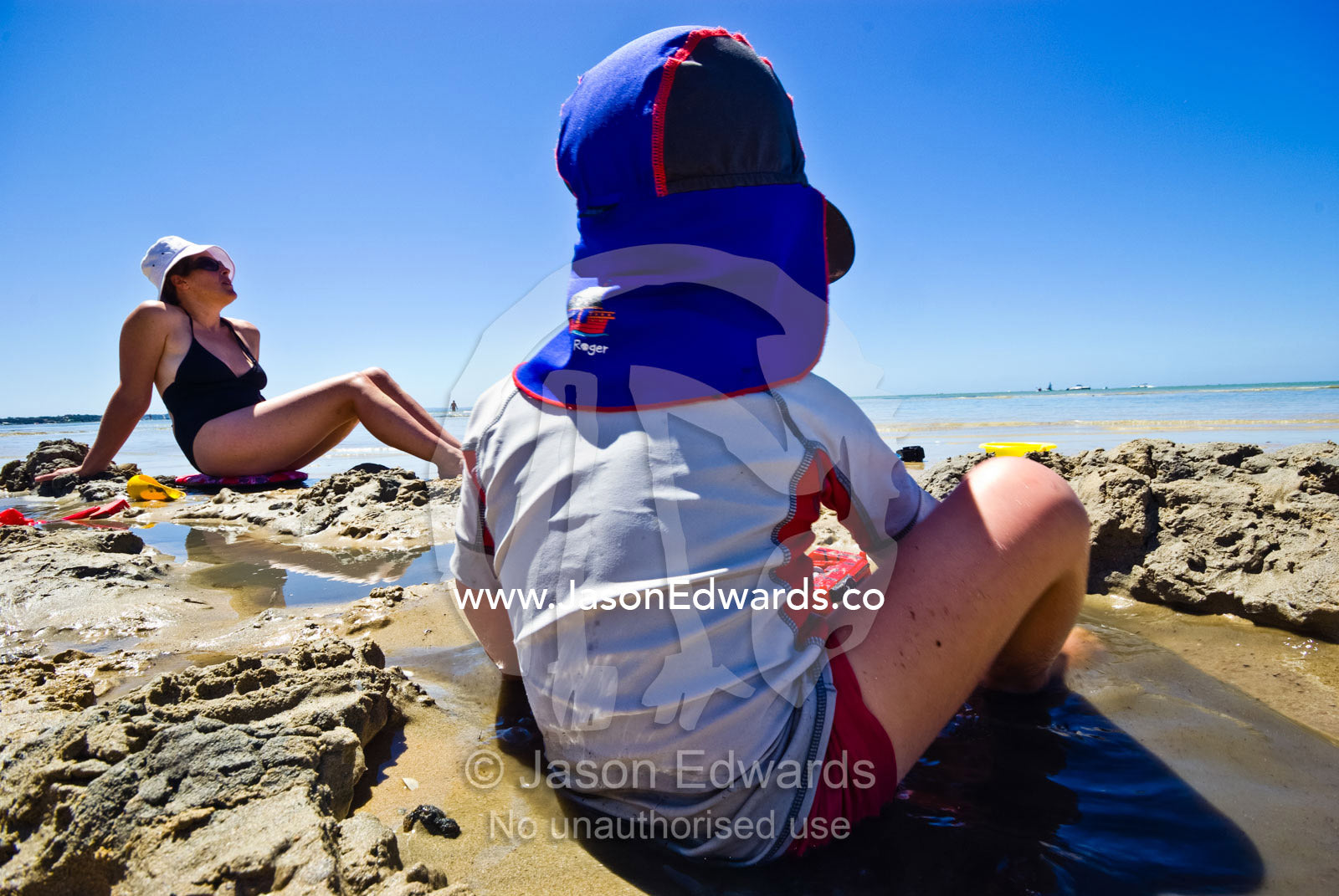 A boy plays in a sandy, muddy puddle on a beach near his mother. Rye Beach, Melbourne, Victoria, Australia.