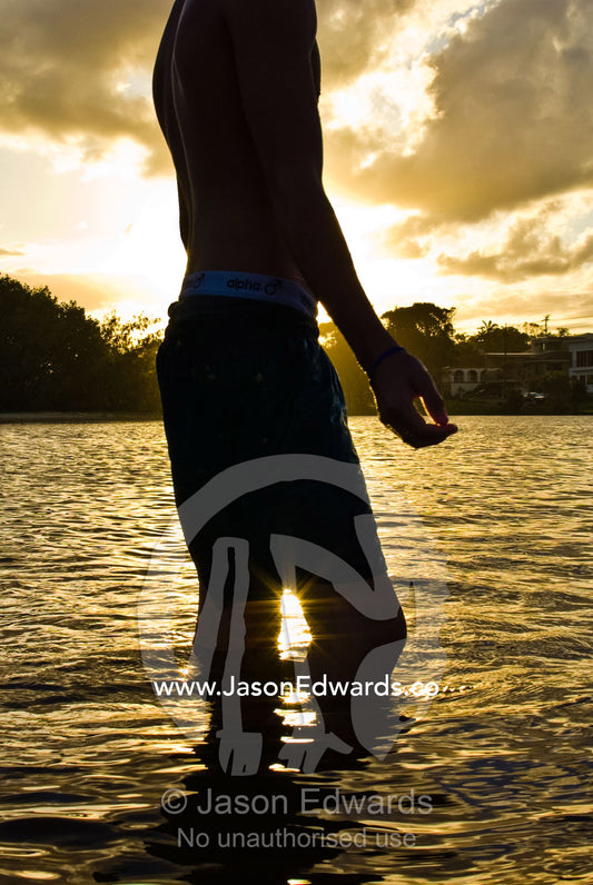 A teenage boy standing in the golden shallows at the beach at sunset. Moffat Beach, Caloundra, Sunshine Coast, Queensland, Australia.