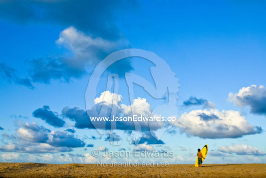 A young boy watches waves with his surfboard on the beach at sunset. Moffat Beach, Caloundra, Sunshine Coast, Queensland, Australia.