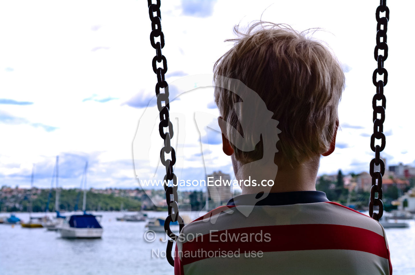A young boy watches boats moored in a bay from a playground swing. East Esplanade Park, Manly Cove, Manly, Sydney Harbour, Sydney, New South Wales, Australia.
