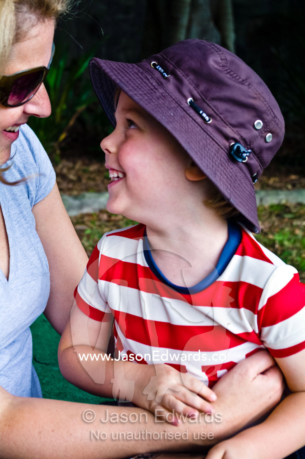 A young smiling boy in a summer hat looks lovingly up at his mother. East Esplanade Park, Manly Cove, Manly, Sydney Harbour, Sydney, New South Wales, Australia.