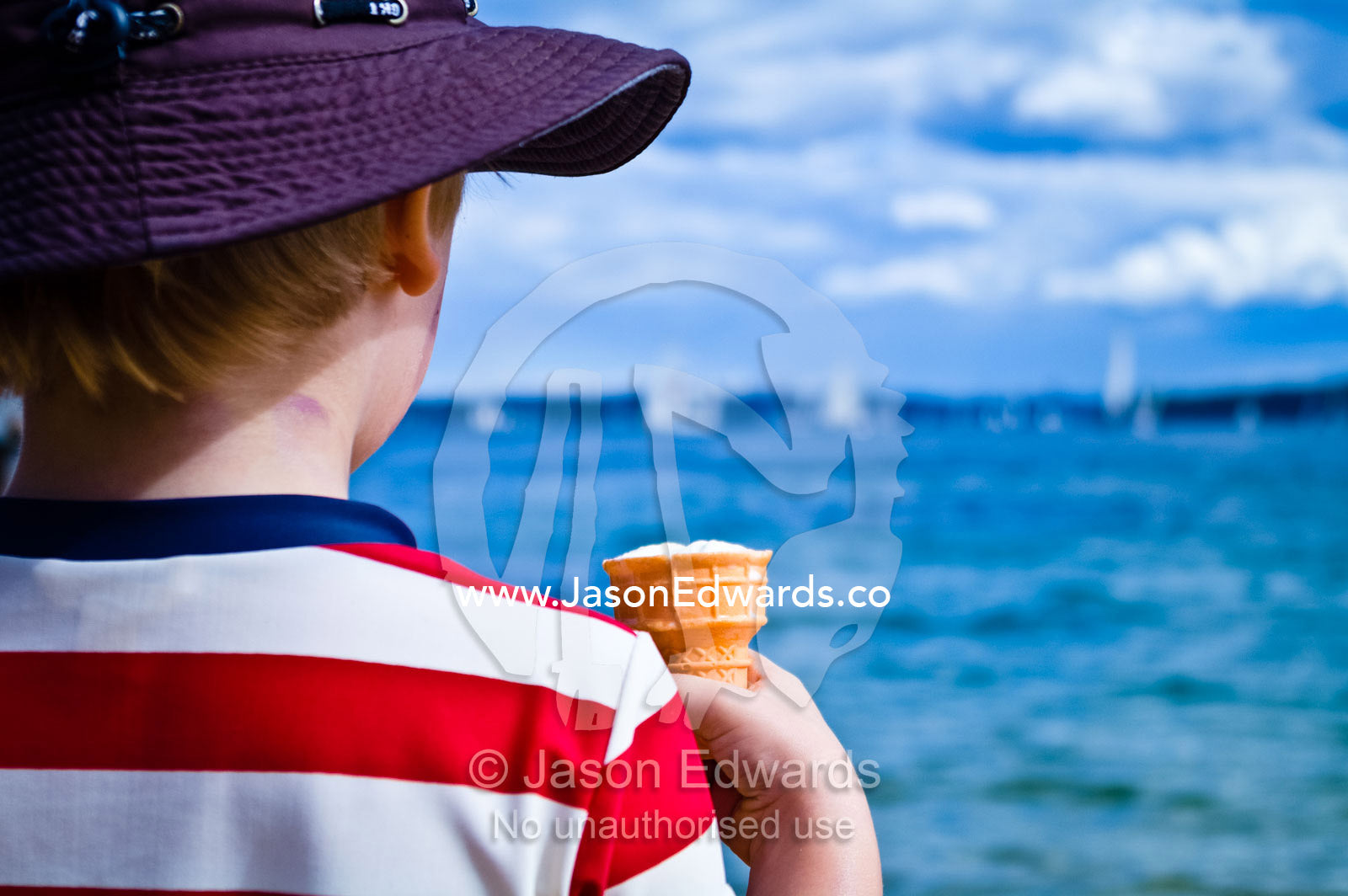 A young boy eats an ice-cream cone whilst watching yachts sailing. Manly Wharf, Manly Cove, Manly, Sydney Harbour, Sydney, New South Wales, Australia.