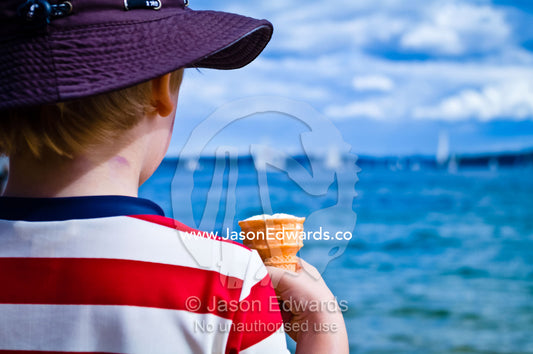 A young boy eats an ice-cream cone whilst watching yachts sailing. Manly Wharf, Manly Cove, Manly, Sydney Harbour, Sydney, New South Wales, Australia.
