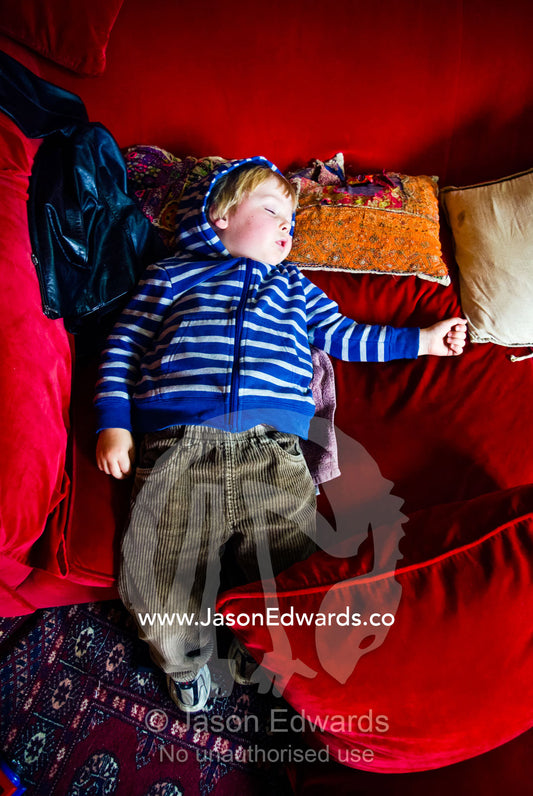 A small boy falling asleep on bright red velvet sofa cushions. Carlton North, Melbourne, Victoria, Australia.