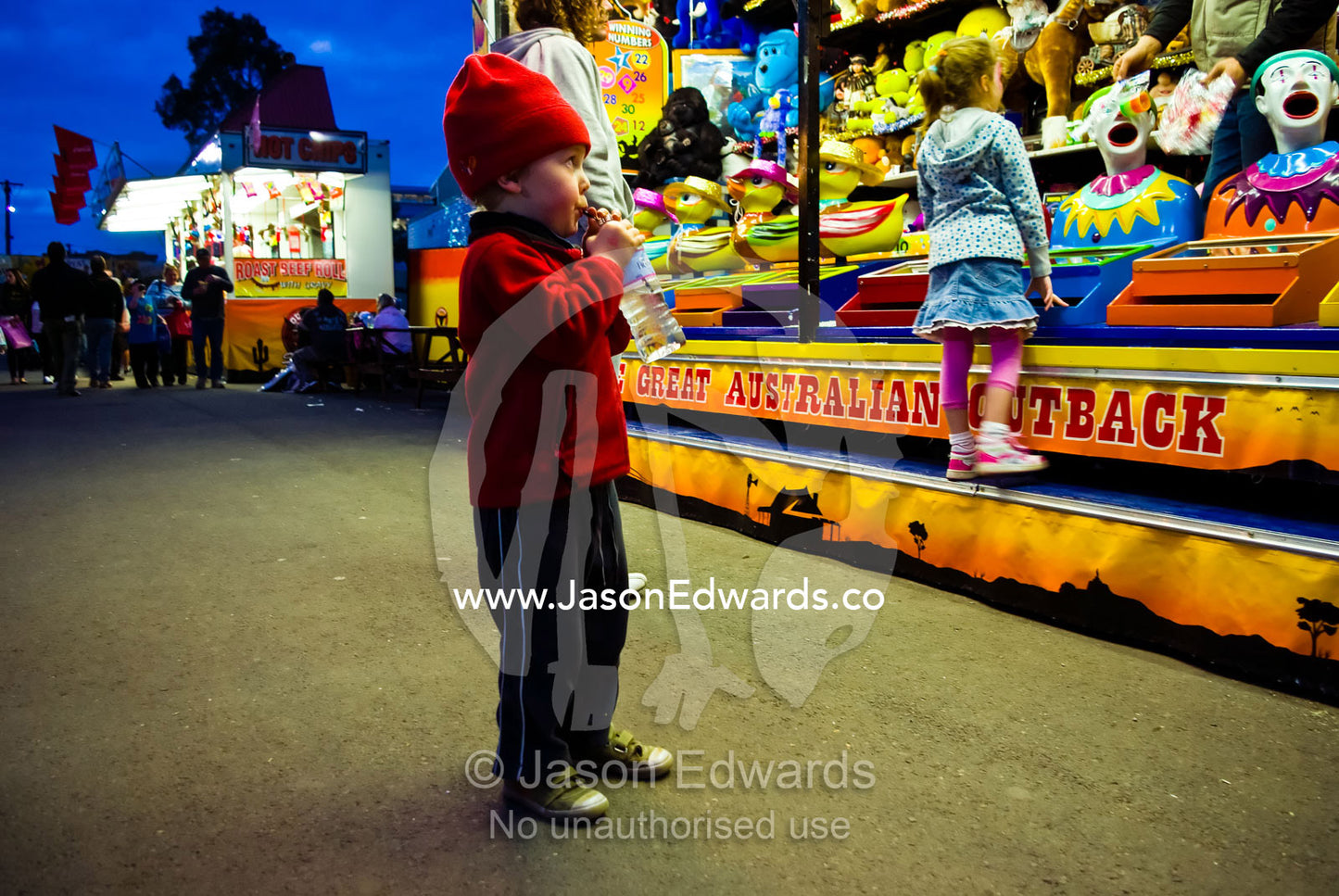 A small boy eats candy and watches a clown game in a fair side show. Royal Melbourne Agricultural Show, Flemington, Melbourne, Victoria, Australia.