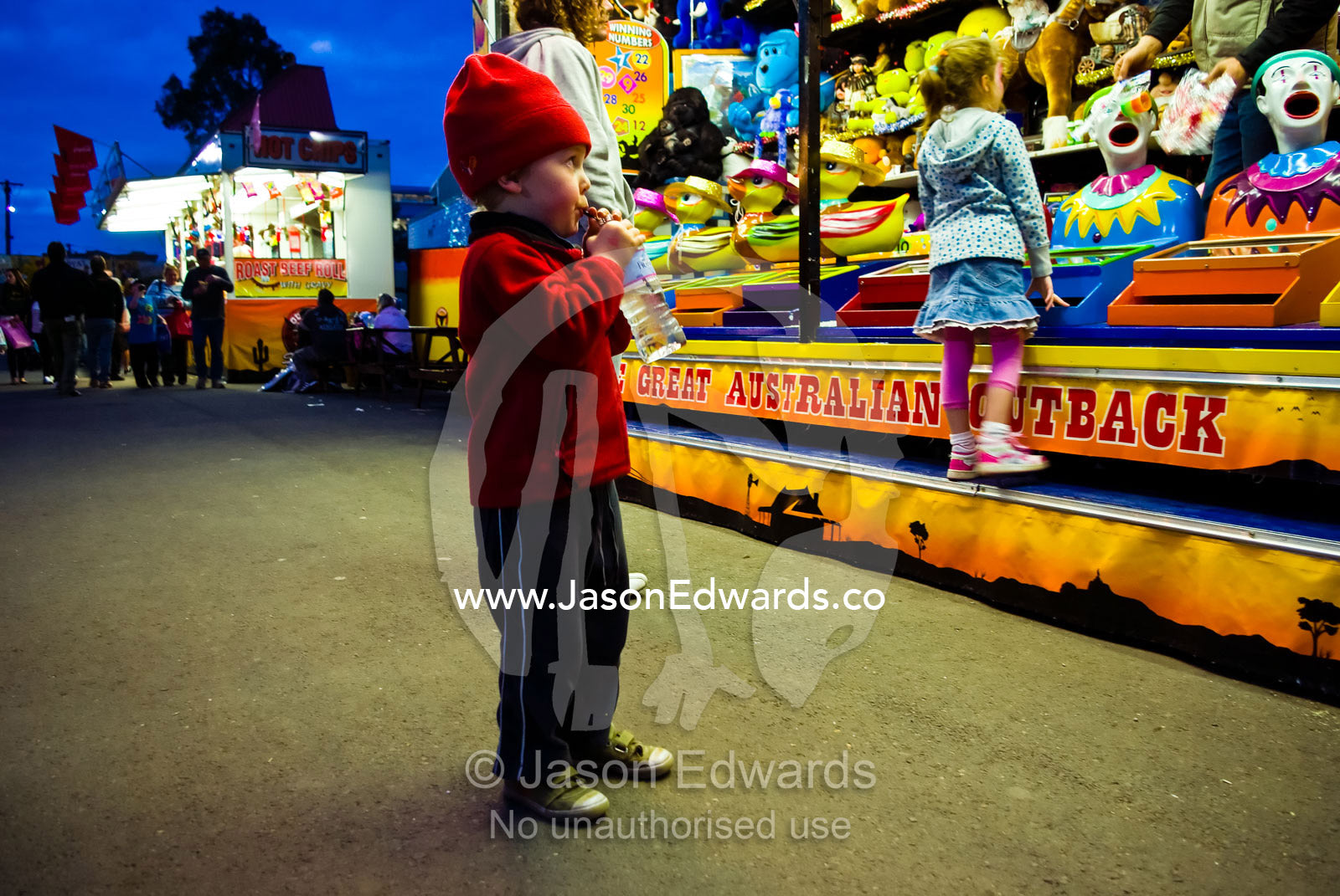 A small boy eats candy and watches a clown game in a fair side show. Royal Melbourne Agricultural Show, Flemington, Melbourne, Victoria, Australia.