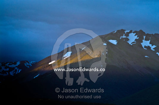 A ray of pastel sun touches a rugged mountain slope above a valley. Ushuaia, Martial Mountains, Fuegian Andes, Ushuaia Bay, The Beagle Channel, Tierra del Fuego Archipelago, Straits of Magellan, Tierra del Fuego Province, Patagonia, Argentina.
