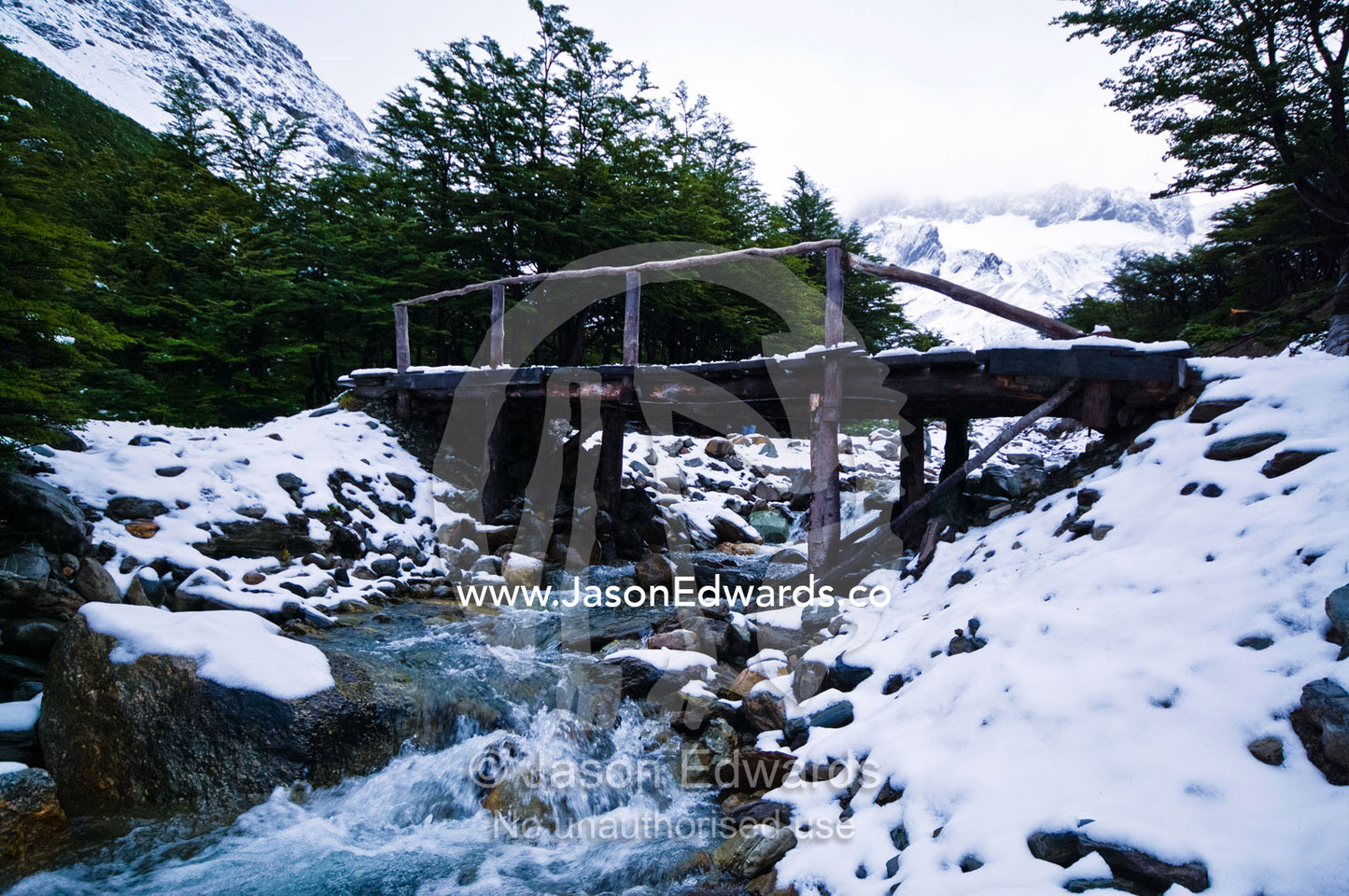 A log and timber footbridge over a stream in a snowy mountain valley. Glacier El Martial, Ushuaia, Fuegian Andes, Tierra del Fuego Archipelago, Straits of Magellan, Tierra del Fuego Province, Patagonia, Argentina.