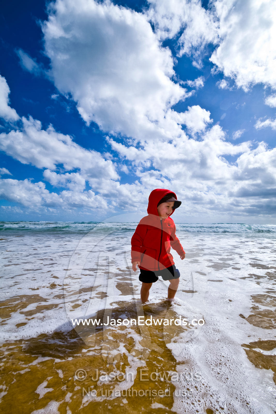 A young boy in a bright red wind jacket wading through the shallows. Venus Bay, Cape Liptrap Coastal Park, Bass Strait, South Gippsland, Victoria, Australia.