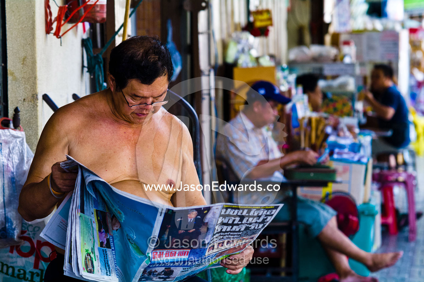 A bare-chested shop owner reads a newspaper waiting for customers. Phra Nakhon District, Rattanakosin District, Bangkok, Thailand.