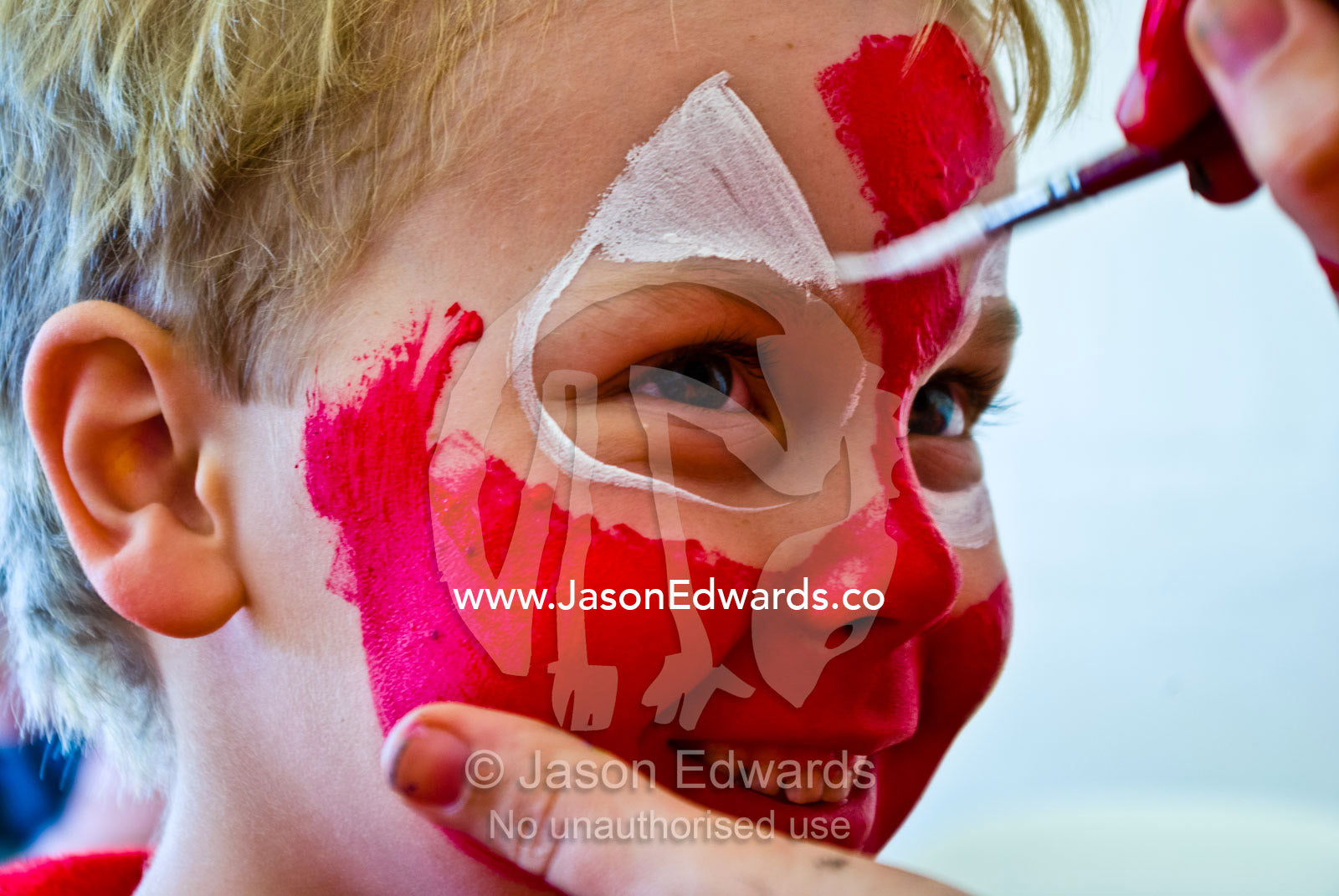 A blonde boy happily has his face painted bright red like spiderman. Frankston, Port Phillip Bay, Melbourne, Victoria, Australia.