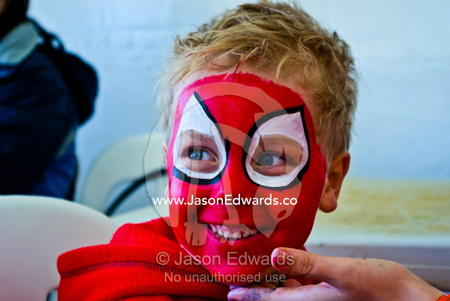 A blonde boy happily has his face painted bright red like spiderman. Frankston, Port Phillip Bay, Melbourne, Victoria, Australia.