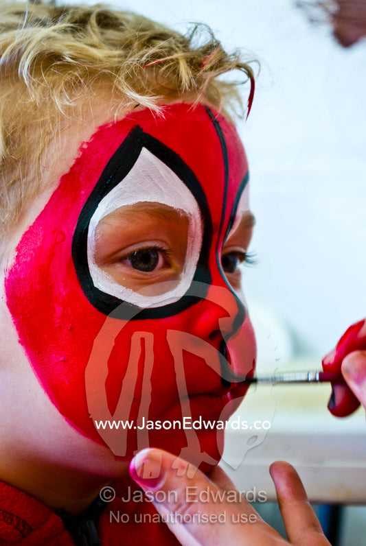 A blonde boy happily has his face painted bright red like spiderman. Frankston, Port Phillip Bay, Melbourne, Victoria, Australia.