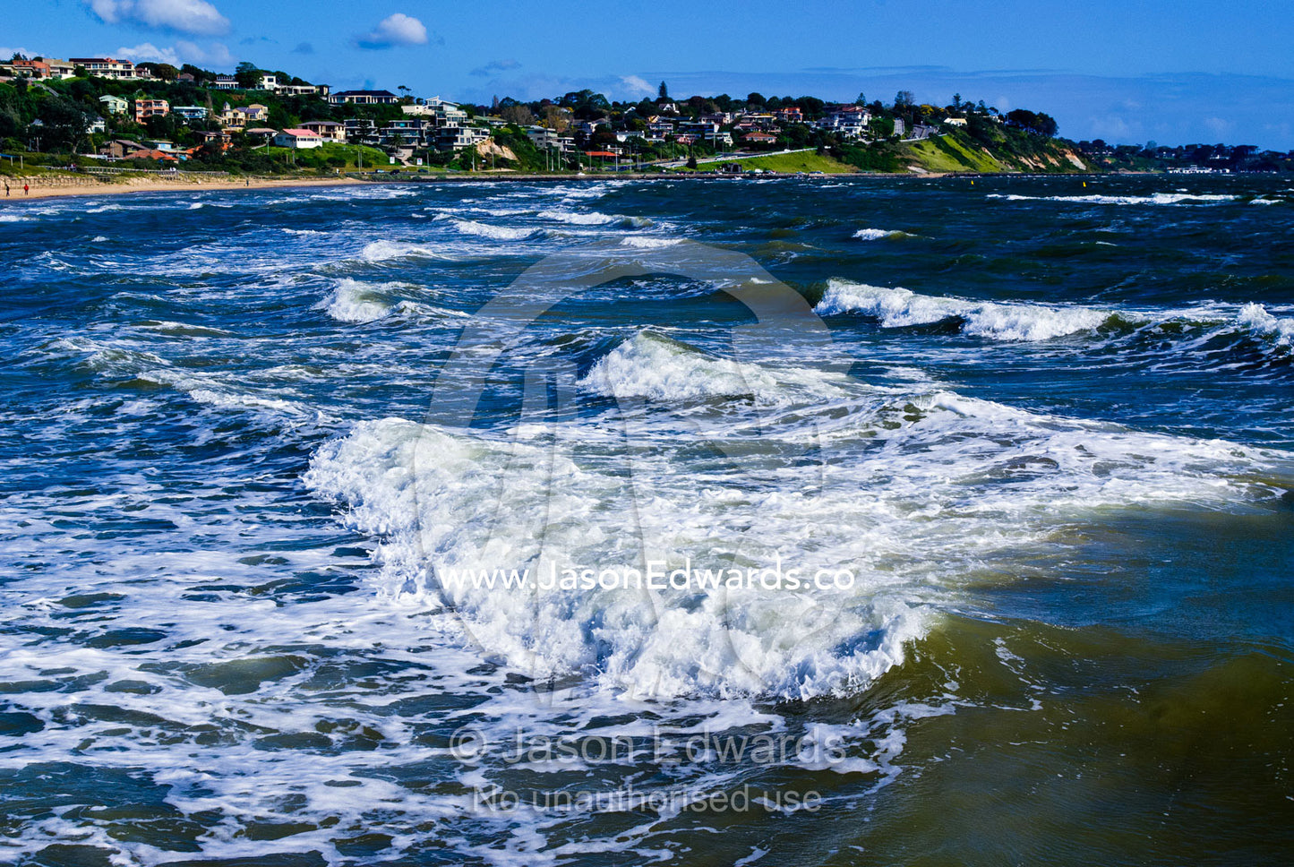 On a windy day foamy waves roll up onto the beach by a coastal suburb. Frankston, Port Phillip Bay, Melbourne, Victoria, Australia.