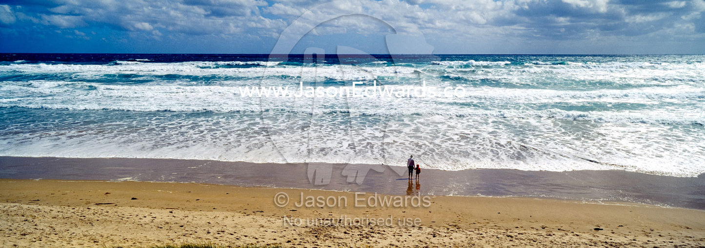A mother and son stand on a wide windswept beach watching foamy waves. Venus Bay, Cape Liptrap Coastal Park, Bass Strait, South Gippsland, Victoria, Australia.
