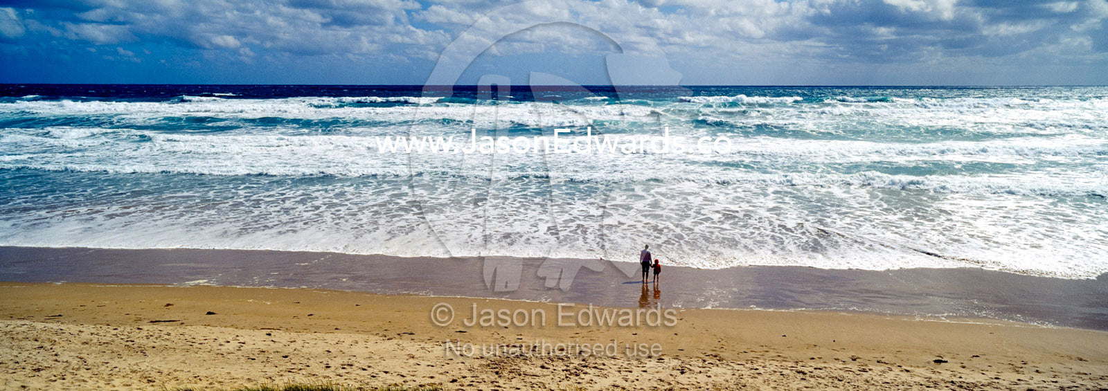 A mother and son stand on a wide windswept beach watching foamy waves. Venus Bay, Cape Liptrap Coastal Park, Bass Strait, South Gippsland, Victoria, Australia.