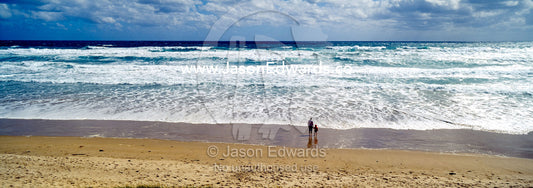 A mother and son stand on a wide windswept beach watching foamy waves. Venus Bay, Cape Liptrap Coastal Park, Bass Strait, South Gippsland, Victoria, Australia.