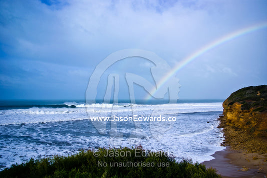 An iridescent rainbow touches the ocean and huge waves at Bells Beach. Bells Beach Surfing Recreation Reserve, Surf Coast, Victoria, Australia.