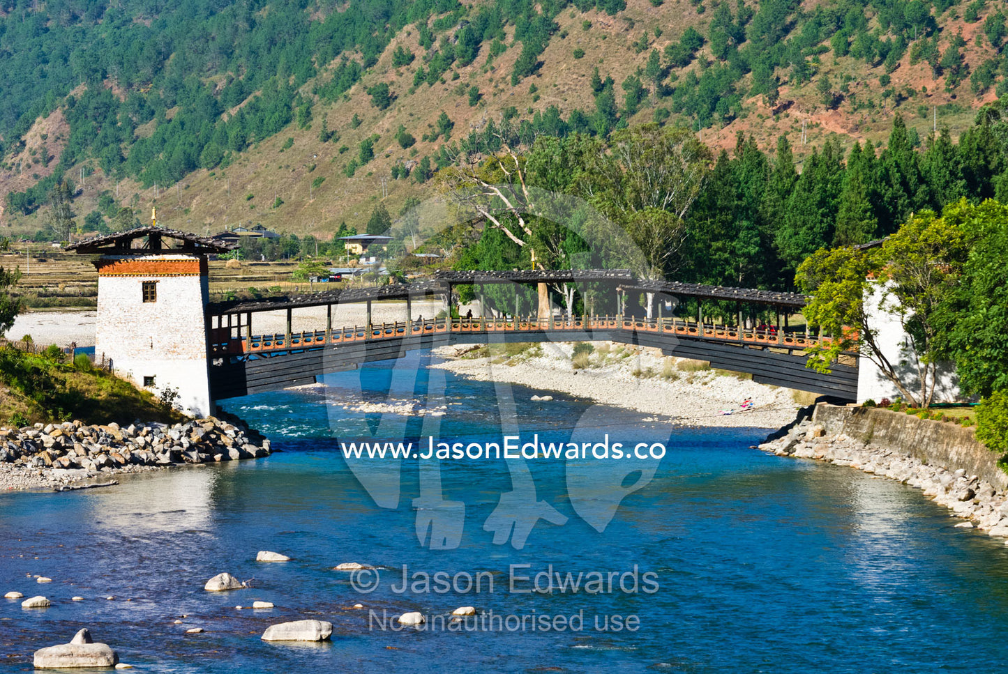 A timber and stone footbridge crosses a turquoise alpine river leading to a Buddhist monastery. Punakha Dzong, Punakha dzongkhag, Punakha District, Bhutan.