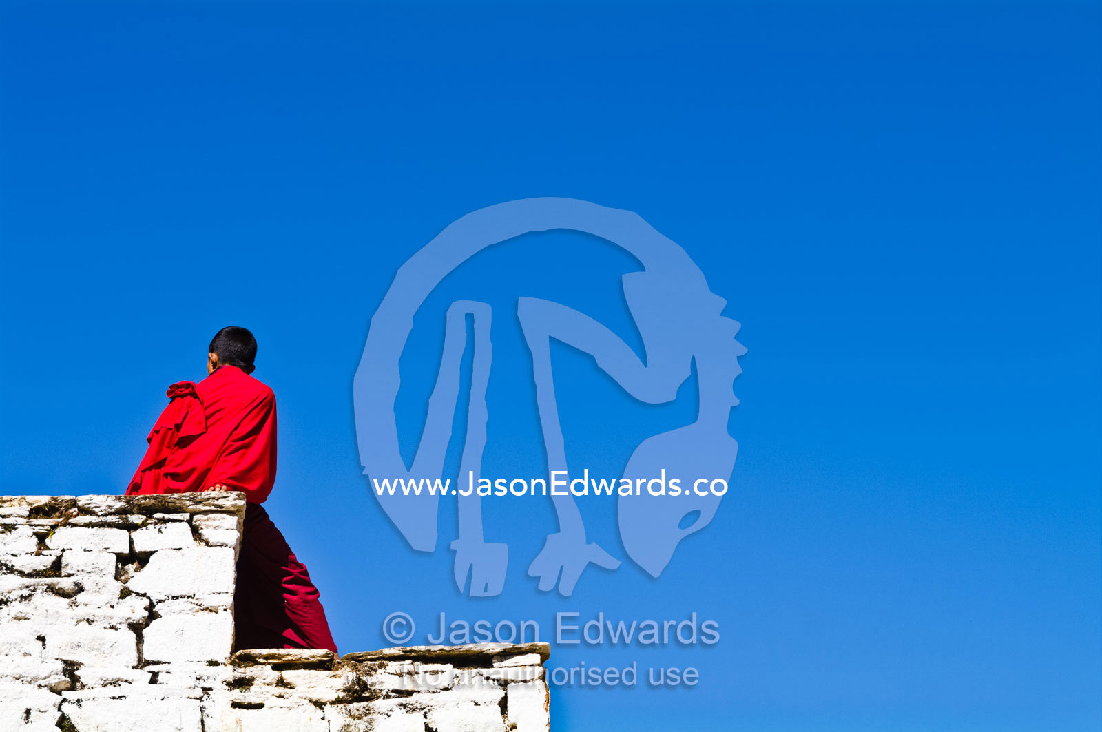 A teenage Buddhist monk sitting in the sunshine on an ancient monastery wall. Rinpung Dzong, Paro Dzong, Paro, Paro District, Bhutan.
