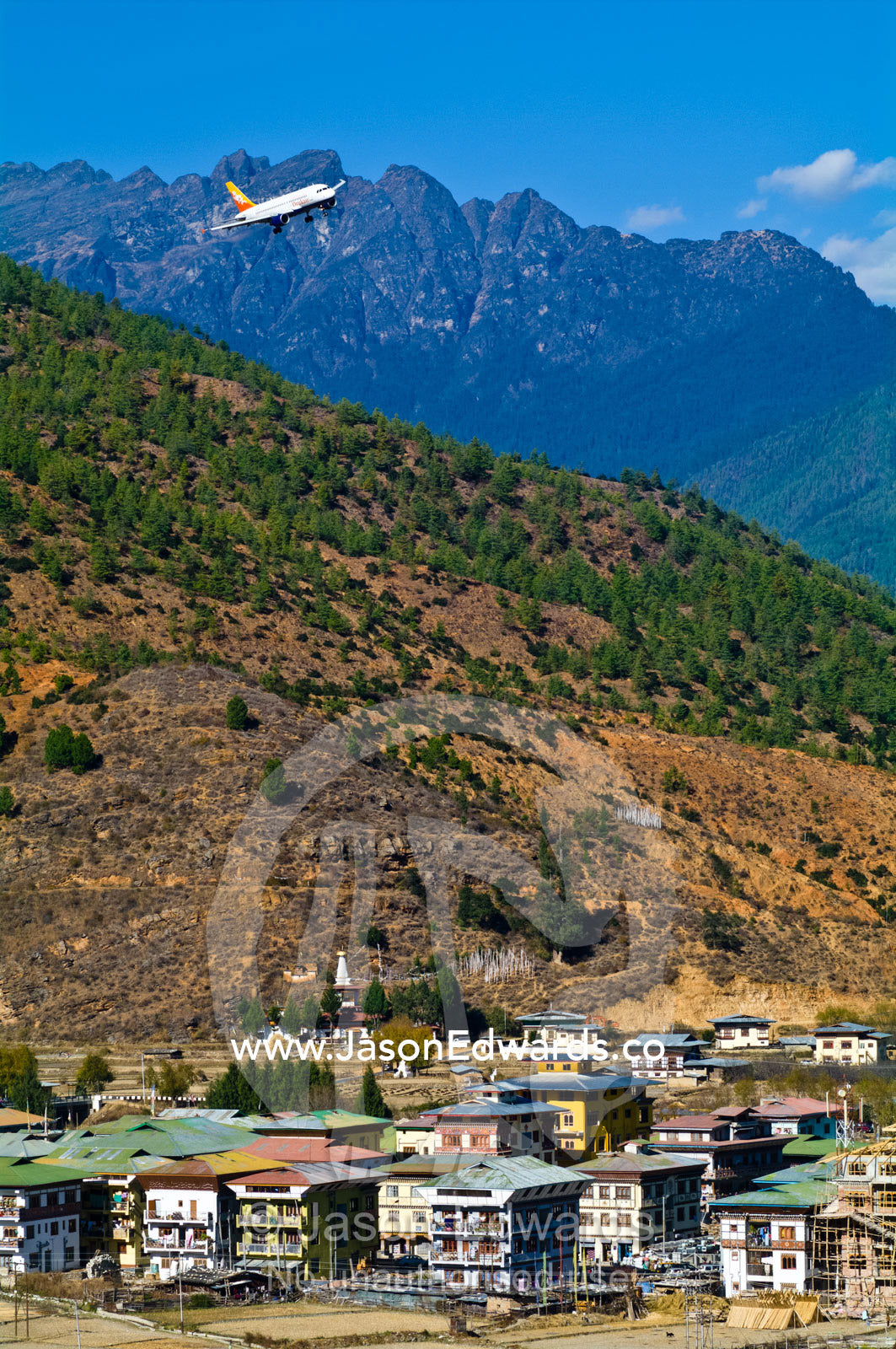 A passenger jet banking over the rooftops of a village in the Himalayan mountains as it begins its landing run. Paro, Paro District, Bhutan.